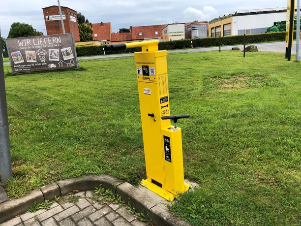 Gelbe Fahrrad-Luftstation am Gehweg, umgeben von Grünflächen und Backsteingebäuden im Hintergrund.