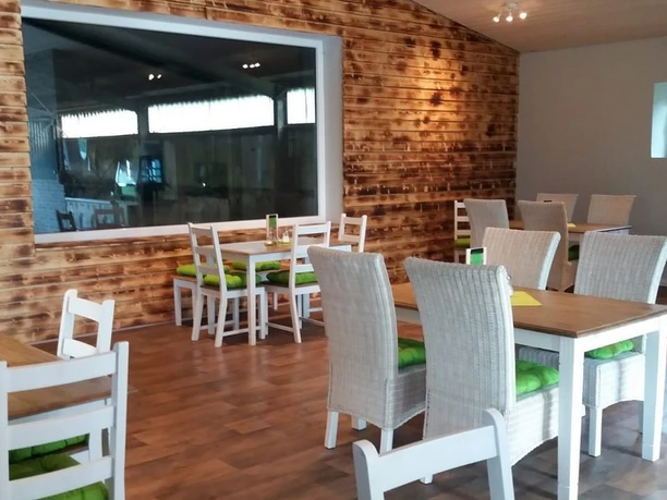 Innenraum eines Restaurants mit weißen Stühlen, grünen Sitzkissen und Holztischen vor einer Wand mit Holzverkleidung