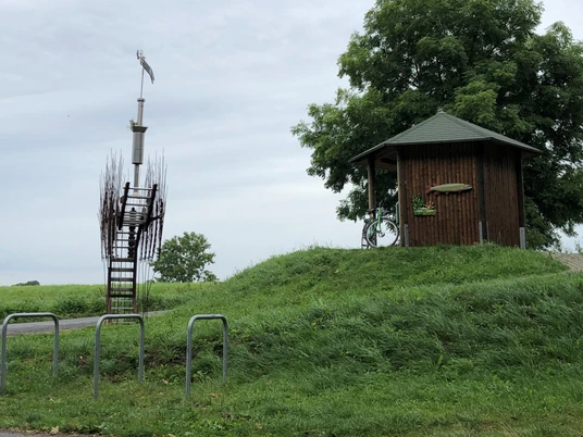 Skulptur mit kunstvoller Treppe und Windspiel, neben hölzernem Pavillon auf grünem Hügel.
