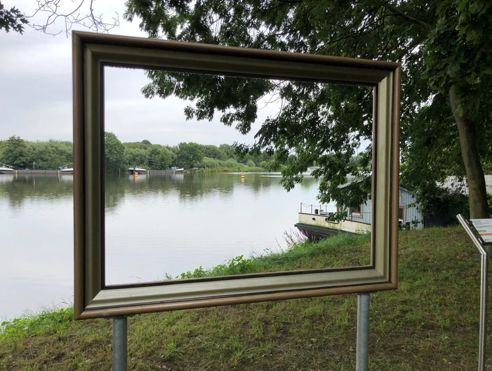 Ein goldener Bilderrahmen steht am Weserufer, mit Blick auf den Fluss und die bewaldeten Uferbereiche.