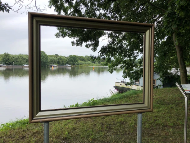 Weserblick 2021 Ein goldener Bilderrahmen steht am Weserufer, mit Blick auf den Fluss und die bewaldeten Uferbereiche.