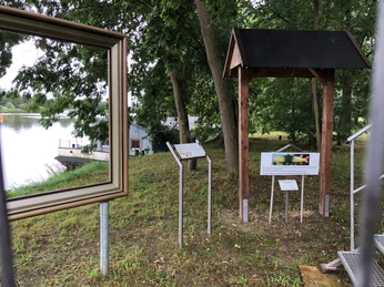 Blick auf eine Holzrahmenstruktur und Infotafeln am Ufer der Weser mit umgebenden grünen Bäumen.