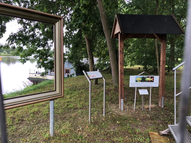 Weserblick 2021 Blick auf eine Holzrahmenstruktur und Infotafeln am Ufer der Weser mit umgebenden grünen Bäumen.