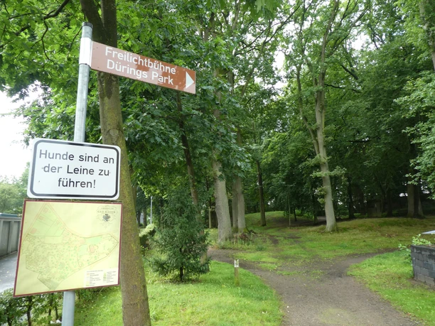 Signpost in Dürings Park pointing to the open-air stage, surrounded by tall, green deciduous trees and a path.