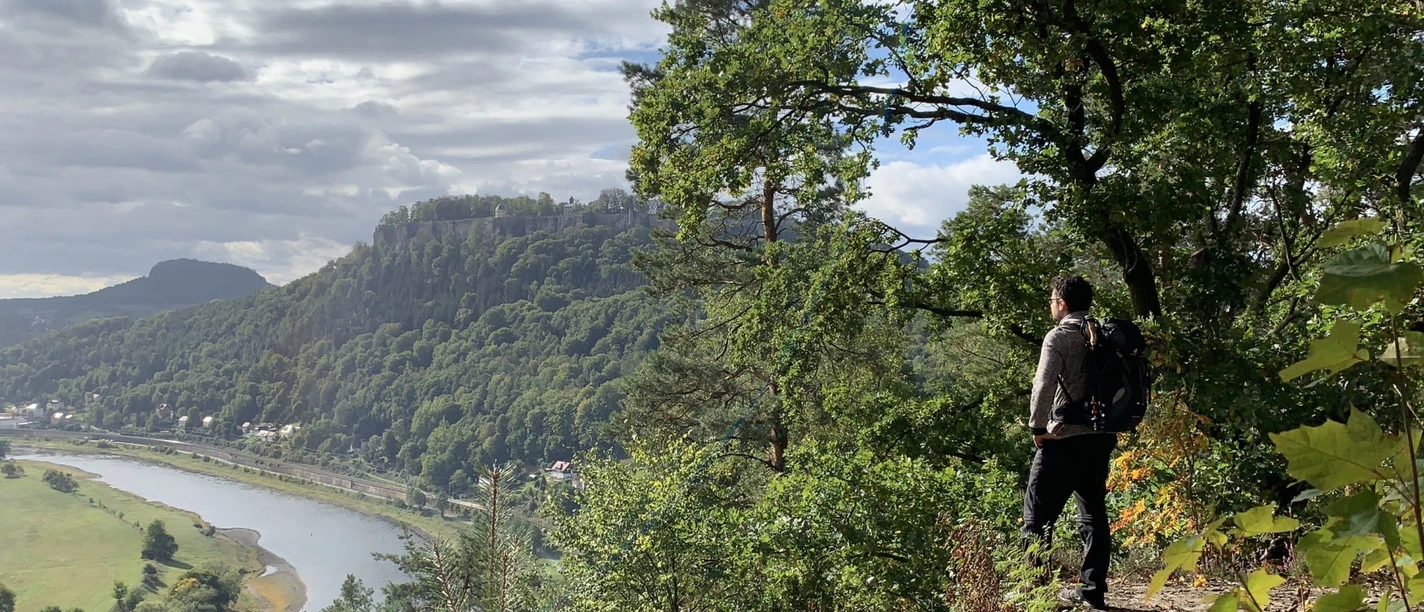 29_Martin-Eichler_Blick-auf-Festung-und-Elbe-vom-Malerweg-aus_Malerwegwegkapelle-2048x1536.jpeg Ein Wanderer blickt von einem bewaldeten Felsvorsprung auf eine Flusslandschaft mit Festung.