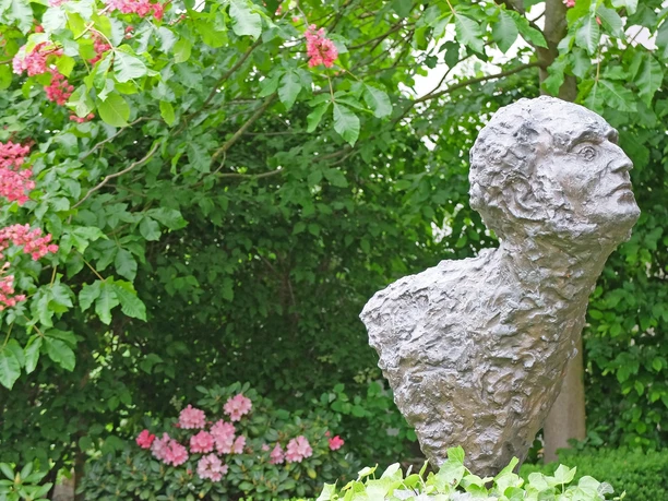 LR_Leipzig_Mendelssohn_Wolfgang Gärtner_DSCF2634.jpg Bronzestatuette eines Mannes im Garten, umgeben von bunten Blüten und grünen Sträuchern.