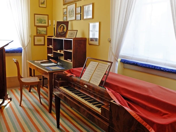 A room with a historic writing desk and piano in warm colors.