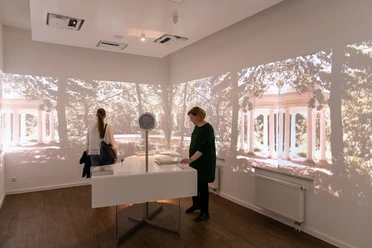 A room with two people and projections of columns and trees on the walls.