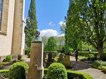 Bronzebüste von Heinrich Hoffmann von Fallersleben auf einem Friedhof, umgeben von grünen Bäumen.