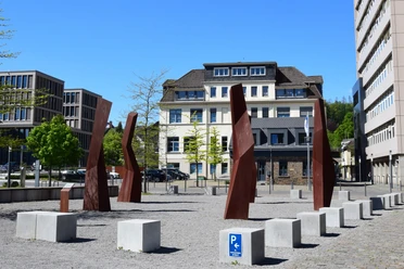 Steinmüllergelände Skulpturen Skulpturen aus rostigem Stahl stehen auf einem Platz in Gummersbach, umgeben von modernen Gebäuden.
