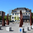 Steinmüllergelände Skulpturen Skulpturen aus rostigem Stahl stehen auf einem Platz in Gummersbach, umgeben von modernen Gebäuden.