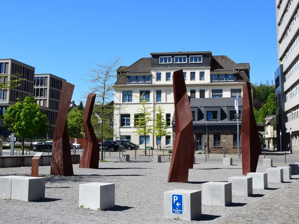 Steinmüllergelände Skulpturen Skulpturen aus rostigem Stahl stehen auf einem Platz in Gummersbach, umgeben von modernen Gebäuden.