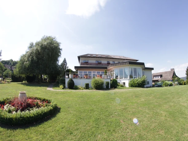 Grüner Rasen vor einem großen, modernen Hotelgebäude mit Terrasse und umliegenden Bäumen.