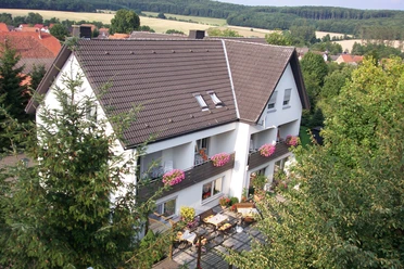Luftaufnahme.jpg Luftbild eines weißen Wohnhauses mit braunem Dach und blühenden Blumenkästen vor Waldkulisse.