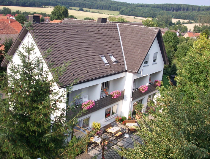 Luftaufnahme.jpg Luftbild eines weißen Wohnhauses mit braunem Dach und blühenden Blumenkästen vor Waldkulisse.