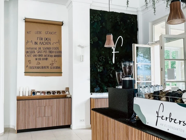 Innenansicht des Cafés mit der Theke rechts, auf der Gläser, Tassen und eine Kaffeemaschine stehen, und kleinen Regal geradeaus, über dem ein Poster mit handschriftlichem Text hängt. Von der Decke hängen zwei Lampen. Die Wände sind hoch.