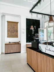 Café Ferbers am Elisengarten Innenansicht des Cafés mit der Theke rechts, auf der Gläser, Tassen und eine Kaffeemaschine stehen, und kleinen Regal geradeaus, über dem ein Poster mit handschriftlichem Text hängt. Von der Decke hängen zwei Lampen. Die Wände sind hoch.