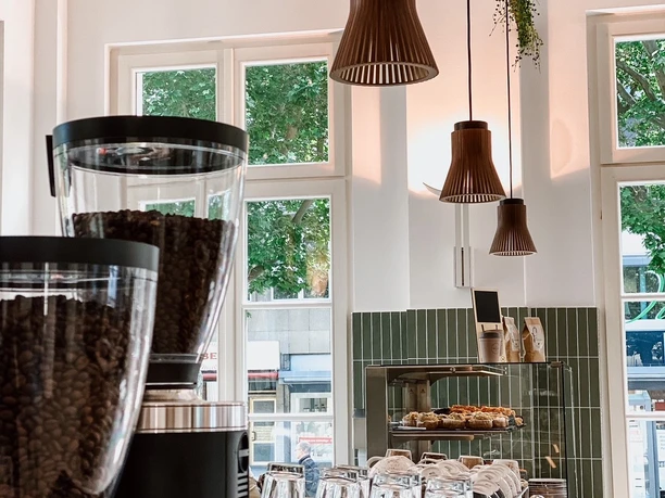 Blick hinter die Theke mit Kaffeemaschine, Gläsern, Tassen, Tellern und Gebäckauslage. Von der Decke hängen mehrere Lampen herab. Durch zwei Fenster kann man auf die Straße und Bäume am Elisenbrunnen schauen.