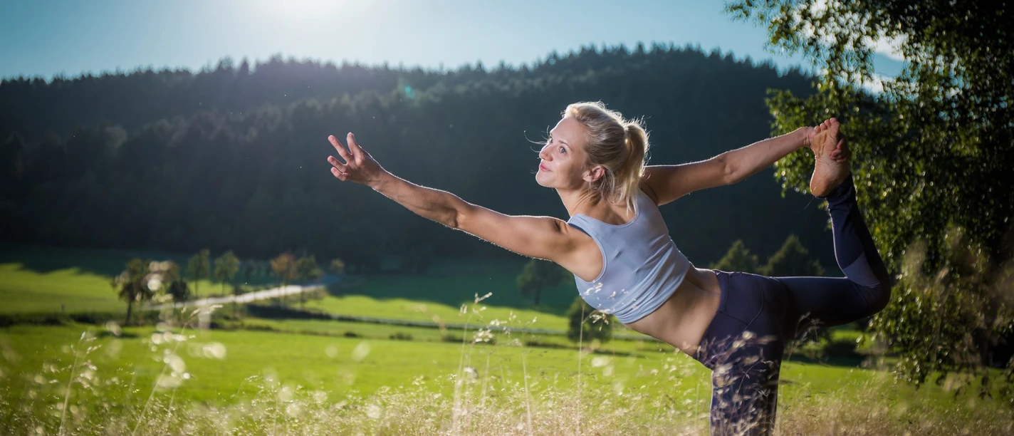 Trixi-Ferienpark_Philipp-Herfort-42.jpg Frau in Yoga-Pose in sonniger Wiese, umgeben von grünen Hügeln und Bäumen.