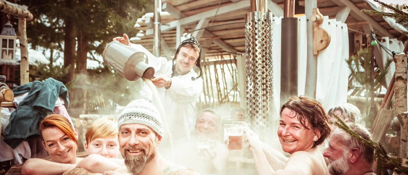Badezuberei-Bierbad-1-Winterdorf-Schmilka-©-Bio-und-Nationalpark-Refugium-Schmilka-Fotograf-Marko-Döhring.jpg Menschen genießen ein warmes Bierbad im Freien in einem hölzernen Hot Tub im Winterdorf Schmilka.