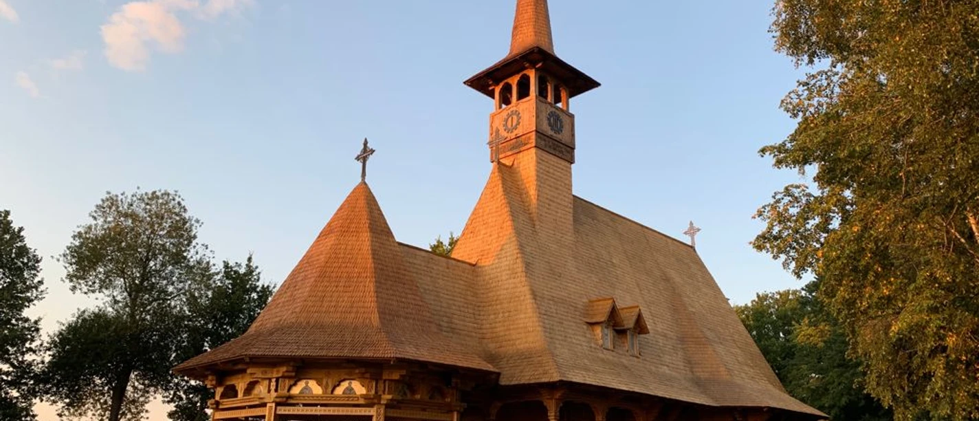 Rumänisch-Orthodoxe-Kirche-Sögel-Christian Lögering.jpg Holzkirche mit spitzem Turm und kunstvollen Schnitzereien im warmen Abendlicht umgeben von Bäumen.