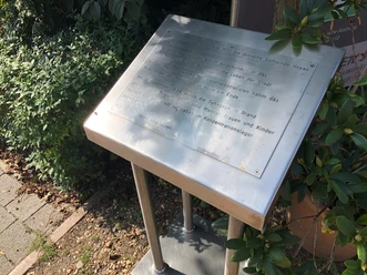 Gedenktafel für die Synagoge Hoya, umrahmt von Pflanzen, auf einem schattigen Platz.