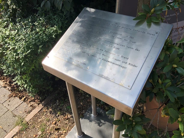 Synagoge Hoya Gedenktafel für die Synagoge Hoya, umrahmt von Pflanzen, auf einem schattigen Platz.