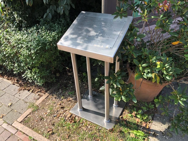 A metal memorial stele with Braille, surrounded by green plants along a paved walkway.