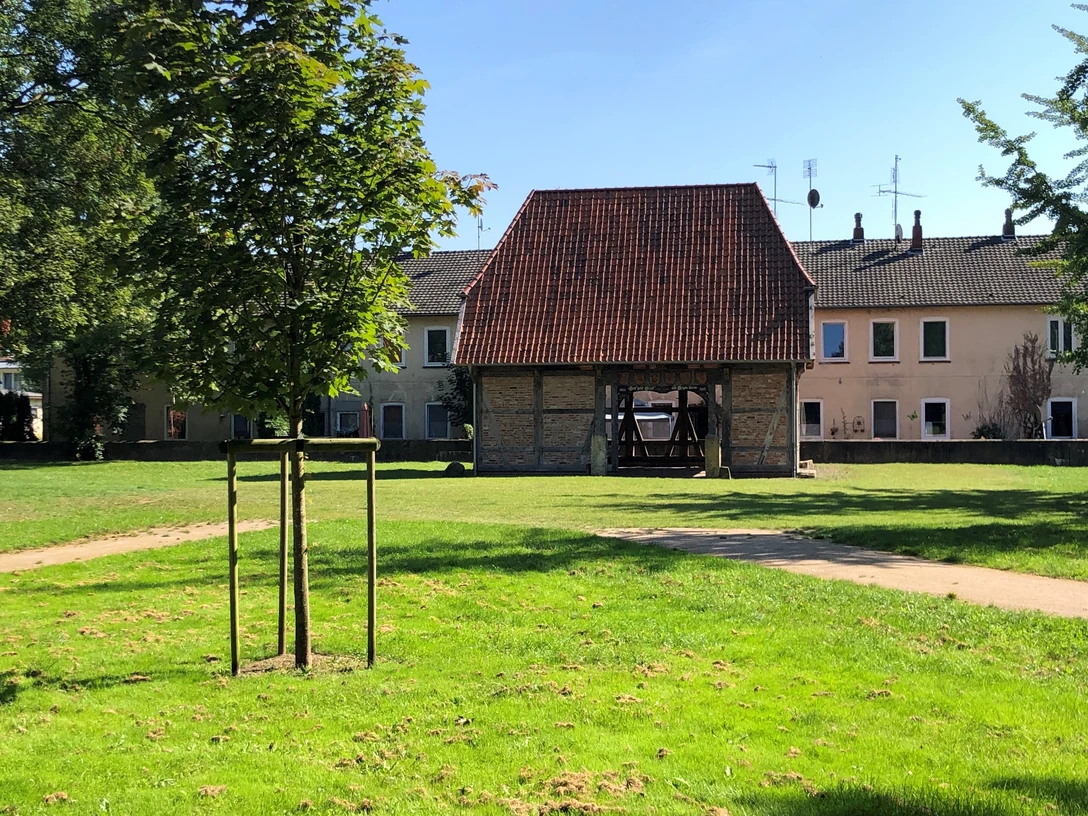 Backsteingebäude mit rotem Ziegeldach und hölzernen Stützen in einer grünen Parklandschaft.