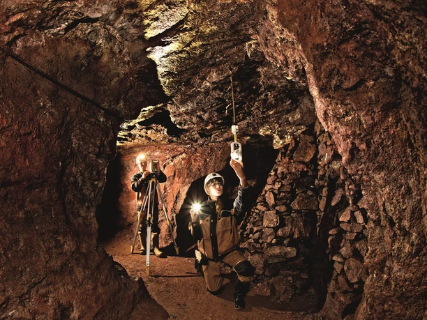 JL121013016_(c)Jürgen Lösel-kleiner.jpg Zwei Menschen erkunden mit Helmen und Lampen eine beleuchtete Höhlenmine aus rotem Gestein.