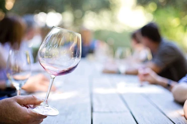 weinglas-am-tisch Das Bild zeigt ein elegant geschwungenes Weinglas, das von einer Hand gehalten wird, während im Hintergrund eine Gruppenversammlung an einem langen Holztisch im Freien stattfindet.