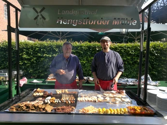 Grillbuffet Das Bild zeigt zwei Männer, die an einem großen Grillbuffet im Freien stehen und verschiedenes Fleisch und Gemüse grillen. Sie lächeln in die Kamera, während köstliche Speisen auf dem Grill brutzeln.