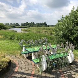 Lieblingsort Alte Staaßenbrücke Es scheint ein Fehler mit der Datei-Suchfunktion zu geben, weshalb ich die Informationen aus der hochgeladenen Datei nicht durchsuchen kann. Dennoch erstelle ich eine allgemeine Bildbeschreibung basierend auf dem bereitgestellten Bild.Gemäldeartige Bänke mit Schilfrohrmotiv an einem gepflasterten Platz, flankiert von grünem Schilf und Blick auf eine Flusslandschaft unter bewölktem Himmel.