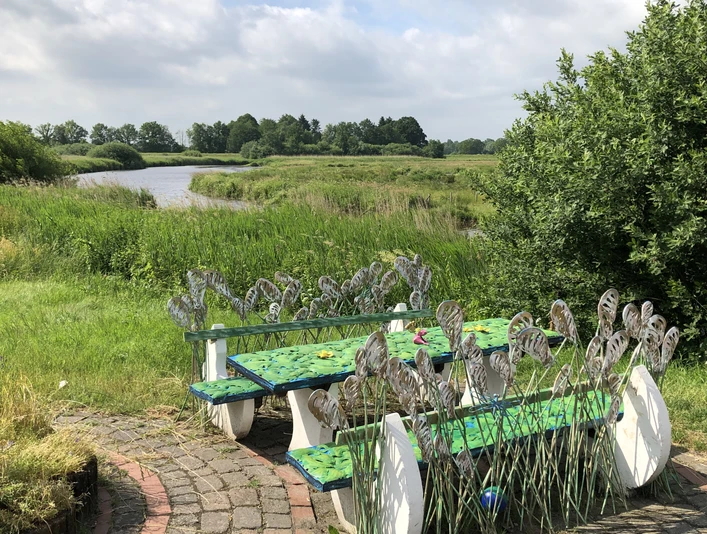 Lieblingsort Alte Staaßenbrücke Es scheint ein Fehler mit der Datei-Suchfunktion zu geben, weshalb ich die Informationen aus der hochgeladenen Datei nicht durchsuchen kann. Dennoch erstelle ich eine allgemeine Bildbeschreibung basierend auf dem bereitgestellten Bild.Gemäldeartige Bänke mit Schilfrohrmotiv an einem gepflasterten Platz, flankiert von grünem Schilf und Blick auf eine Flusslandschaft unter bewölktem Himmel.