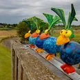 Bunte Vogelschar am Deich Eine Reihe bunter Vogel-Skulpturen auf einem Geländer, mit einer grünen Landschaft im Hintergrund.