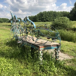 Lieblingsort Heeren-Wehren-Brücke Eine kunstvoll verzierte Bank steht auf einer Wiese, umgeben von üppigem Grün und einem blauen Himmel im Hintergrund. Die geschwungenen Metallverzierungen sind in bunten Farben gehalten und verleihen der Szenerie eine künstlerische Note.