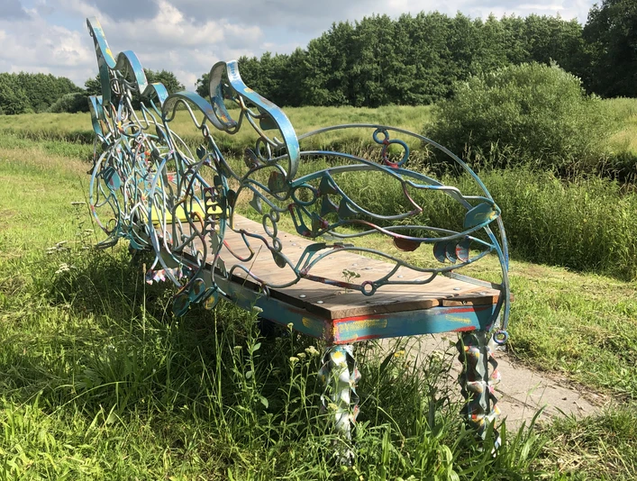Lieblingsort Heeren-Wehren-Brücke Eine kunstvoll verzierte Bank steht auf einer Wiese, umgeben von üppigem Grün und einem blauen Himmel im Hintergrund. Die geschwungenen Metallverzierungen sind in bunten Farben gehalten und verleihen der Szenerie eine künstlerische Note.