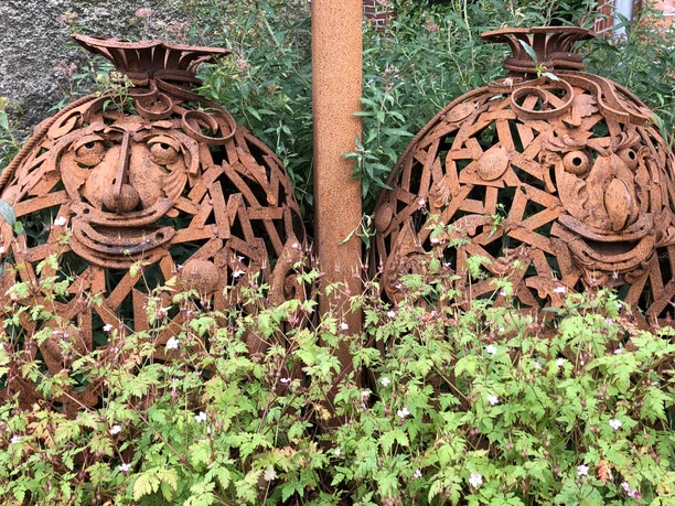 Mehlsäcke - Lieblingsort Hengstforder Mühle Zwei Mehlsäcke aus Stahl, verziert mit lächelnden Gesichtern, eingebettet in üppiges Grün.