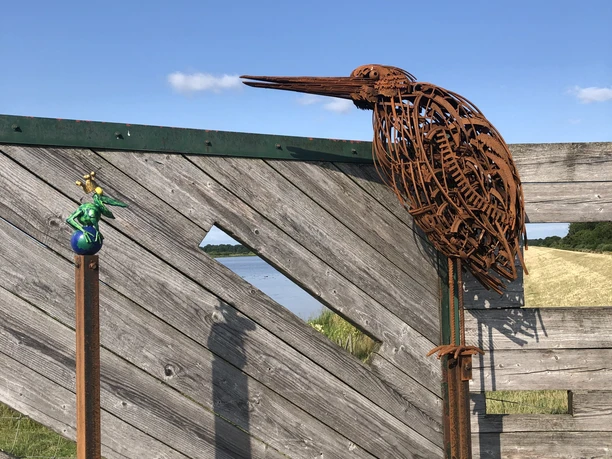 Reiher und Frosch Das Bild zeigt eine stilisierte Reiherfigur aus rostfarbenen Stahll, die auf einem Holzzaun sitzt. Daneben ist ein kleiner, bunt gestalteter Frosch dargestellt, der ebenfalls auf einer Metallstange befestigt ist. Der Hintergrund besteht aus blauem Himmel und grüner Landschaft.