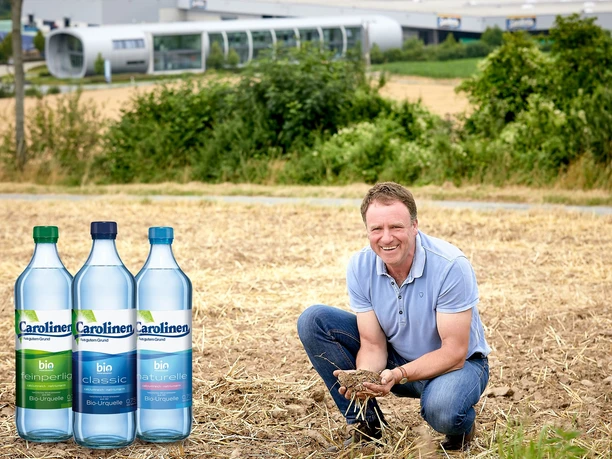 Carolinen Mineralbrunnen - Bio-Landwirt Bobbert Ein lächelnder Landwirt in einem Feld mit Carolinen-Wasserflaschen im Vordergrund und einem modernen Gebäude im Hintergrund.