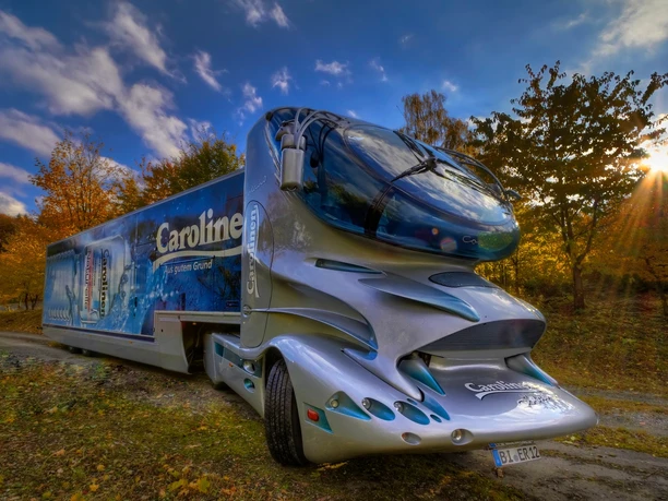 Carolinen Mineralbrunnen - Colani-Truck Futuristischer Carolinen Mineralbrunnen-Truck, umgeben von herbstlichem Wald, bei strahlendem Sonnenschein.