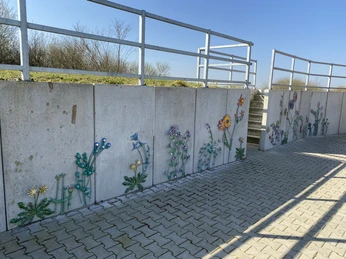 Blumenwand am Deich Eine Betonmauer am Deich ist kunstvoll mit bunten, stilisierten Metallblumen verziert.