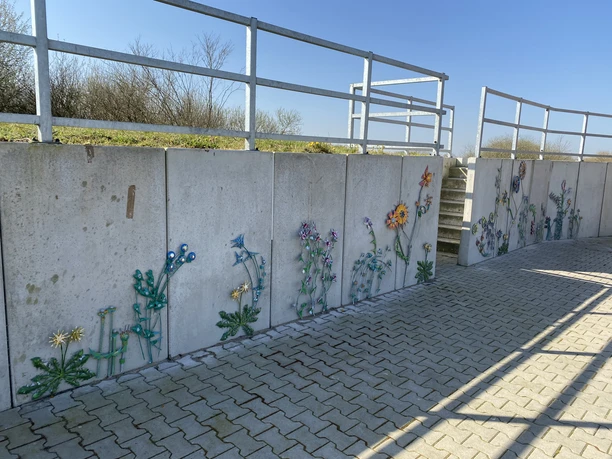 Blumenwand am Deich Eine Betonmauer am Deich ist kunstvoll mit bunten, stilisierten Metallblumen verziert.