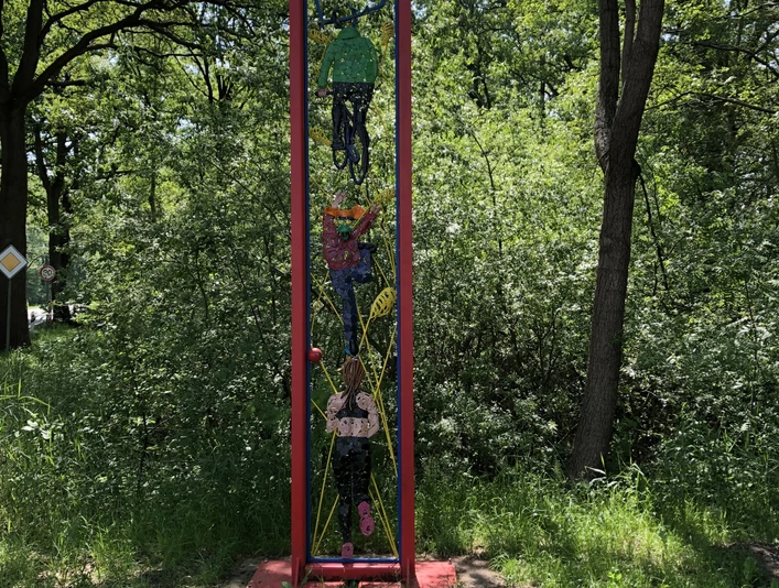 Lieblingsort Unterster Weg Schmale rote Metallskulptur mit abstrakten Formen, vor dichtem grünem Wald aufgestellt.