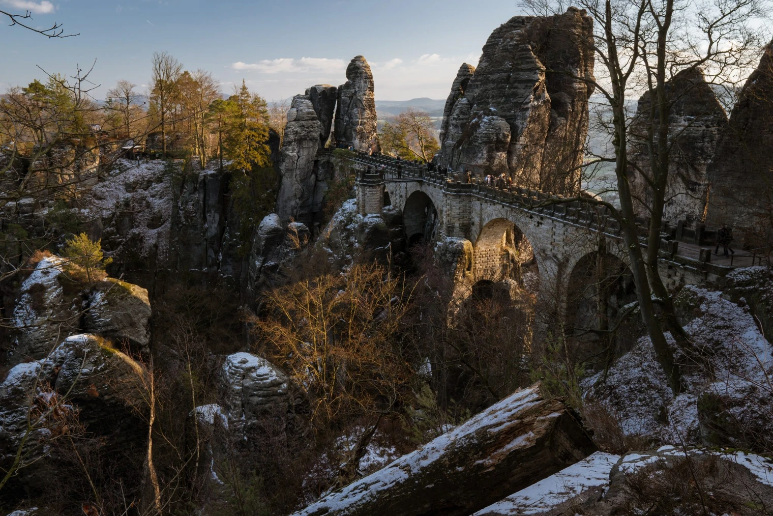 Tour-24-Großer-Nordic-Walking-Bastei-Trail-8.jpg Natursteinbrücke im Licht der Abendsonne, umgeben von steilen Felsen und kahlen Bäumen.