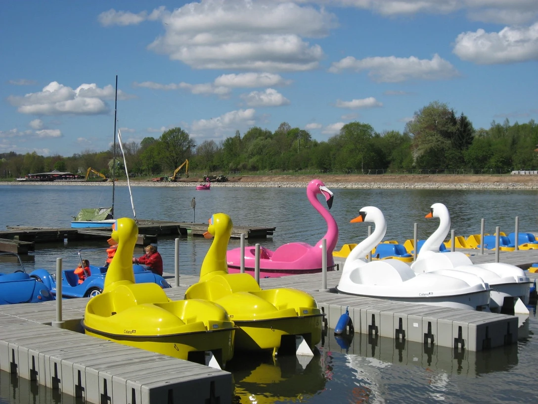 Tretboote schwimmen auf einem See, darunter gelbe Enten- und weiße Schwanboote unter blauem Himmel.