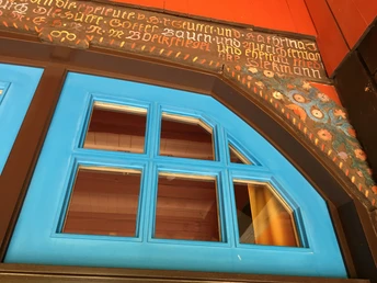 Ausschnitt Künstlerhaus - Böckstiegel Detailreiche, blaue Holzfenster mit antiker Inschrift und floralen Verzierungen auf einem historischen Gebäude.