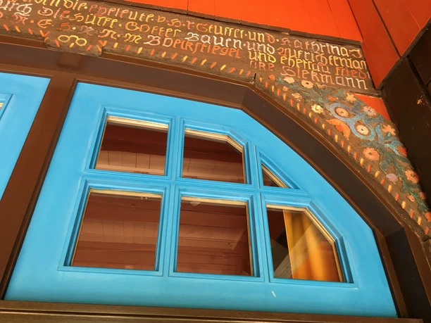 Ausschnitt Künstlerhaus - Böckstiegel Detailreiche, blaue Holzfenster mit antiker Inschrift und floralen Verzierungen auf einem historischen Gebäude.