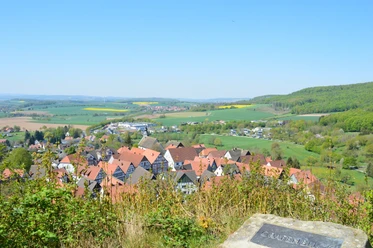 Blick auf ein Dorf mit roten Dächern, umgeben von grünen Hügeln und Feldern unter klarem Himmel.