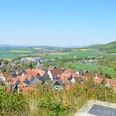 Blick auf ein Dorf mit roten Dächern, umgeben von grünen Hügeln und Feldern unter klarem Himmel.